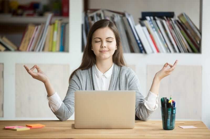 Female student sitting at a desk with a laptop in front of her. She has her eyes closed and has her hands in a meditative pose