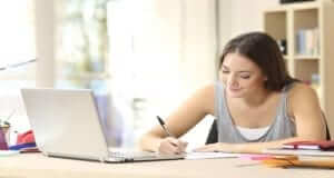 Young brunette woman sitting at a desk writing. Desk has a laptop, post-it notes, and books. There is a bookcase in the background.