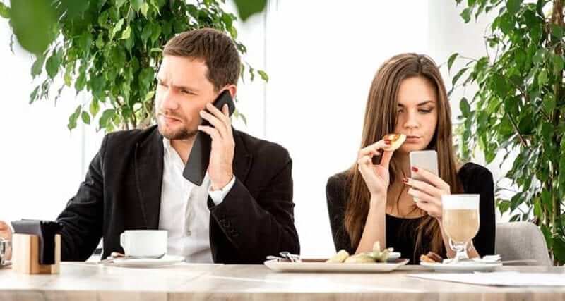 Male and female sitting at a lunch table. Male is talking on the phone and female is looking at her phone.