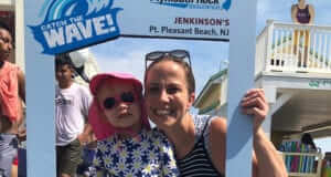 a mom and baby daughter smiling and wearing sunglasses peeking through a teacher week at the beach sign for photos at jenkinsons boardwalk