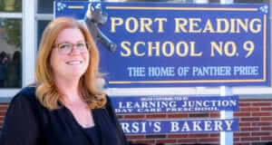 Tricia Fitzgerald, Principal in Woodbridge Township School District, has red hair, freckles, and glasses and is smiling standing next to the school sign that reads "Port Reading School No. 9 The Home of Panther Pride"