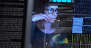 Reflection of woman with glasses and holding pencil near mouth looking at computer screen which is filled with small font of numbers, letters, codes, and the globe