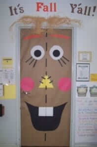 Classroom door decorated as a buck toothed scarecrow. At the top of the door reads "It's Fall Y'all!"