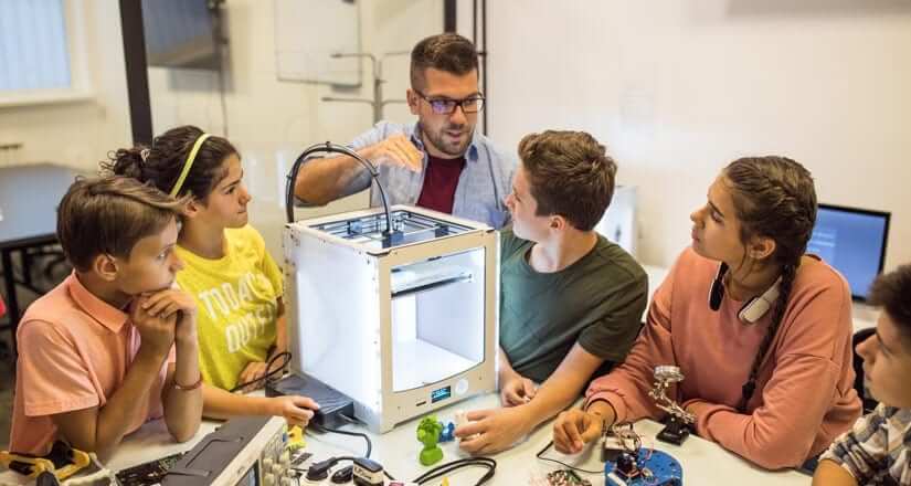 Male teacher with glasses and short sleeve button down shirt presenting 3-D printing to students in classroom