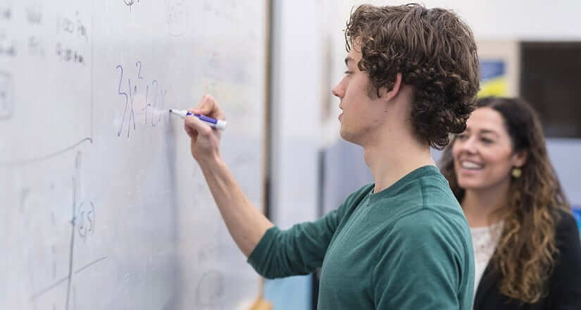 Male high school student writing a math equation on a white board with female teacher with long, brown curly hair standing next to him