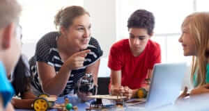 A female teacher and students standing around a desk with a laptop and parts to build a remote control car