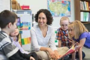 Female teacher with short, dark, curly hair sitting on the floor reading a book to young students in the classroom