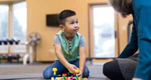A teacher sitting on the floor of a classroom talking to a young student who is playing with blocks