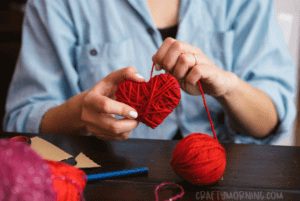 Hands of a female with her sleeves rolled up making a heart out of red yarn