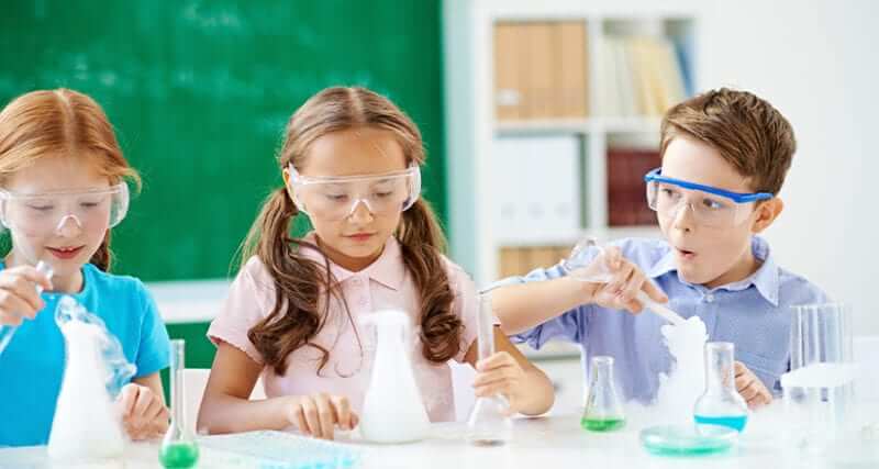 Three children in a chemistry classroom wearing goggles and working with Erlenmeyer flasks, florence flasks, test tubes, and creating chemical reactions