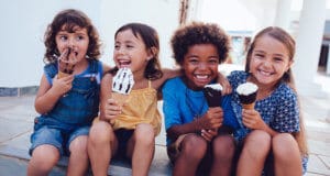 Four children sitting on a curb eating ice cream cones with their arms wrapped around each other smiling and laughing