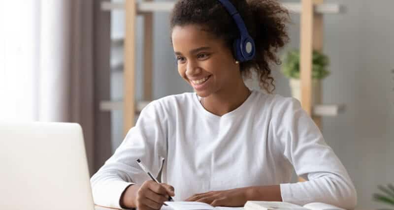 African American female student wearing headphones, holding a pen and notebook, looking at a laptop and smiling