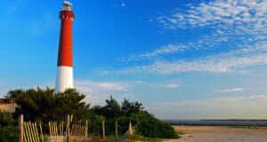 A white and red lighthouse with blue skies in the background and sand and a dunes fence
