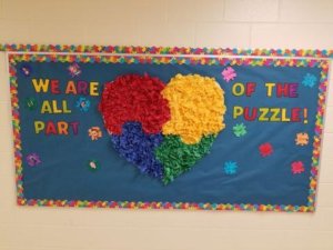 A bulletin board that says "We are all part of the puzzle". The background is blue. The boarder is puzzle pieces. In the center is puzzle of a heart made into 4 pieces that are red, yellow, blue and green