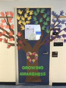 A classroom door decorated as a tree. The leaves were made by tracing students' hands on construction paper and cutting them out. Near the trunk of the tree it says "Growing Awareness"
