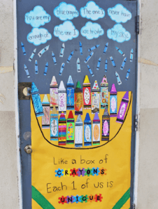 A classroom door that says "Like a box of crayons, each 1 of us is unique". There is an image of a box of crayons. The crayons were decorated by the students. There are cut outs of clouds at the top of the door.