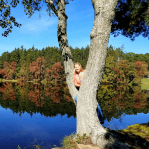 Jessica Cicalese Kurtz wearing a lanyard leaning again a tree. A lake and trees with autumn leaves in the background.