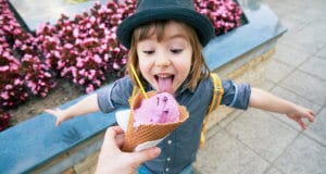 A hand holding an ice cream cone out to a child who has his tongue out about to lick it