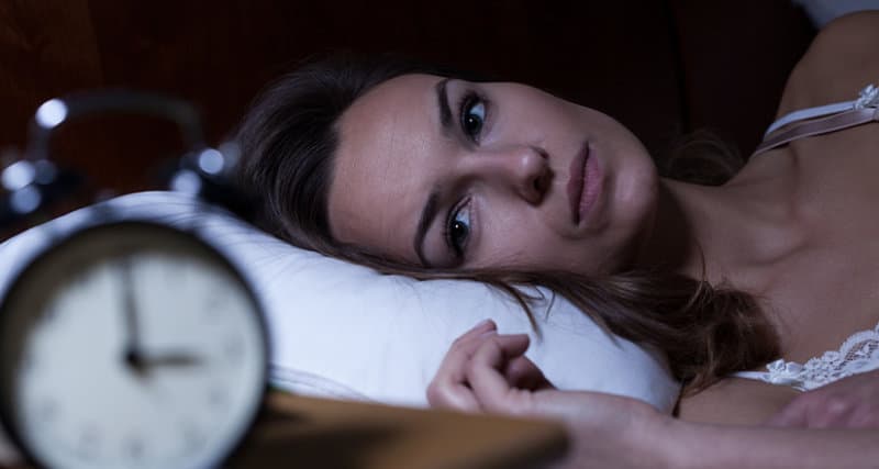 A woman laying in bed looking at an alarm clock on a night stand