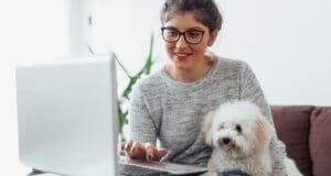 A female wearing glasses and touching the keypad on a laptop with a small dog sitting with her