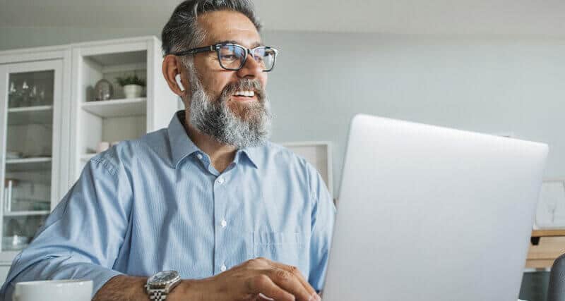 An older gentleman with gray hair and a gray beard wearing glasses and Apple EarPods siting in front of a laptop smiling