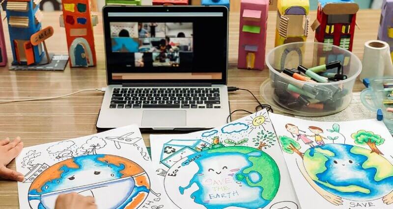A laptop showing a virtual classroom in session surrounded by school supplies and artwork