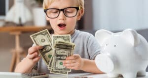 Young blonde boy with glasses looking surprised as he counts cash in his hands with a piggy bank next to him