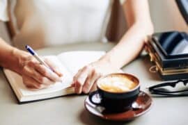 person sitting at a table writing daily intentions in journal with cup of coffee