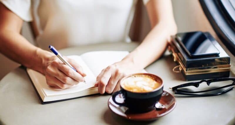 person sitting at a table writing daily intentions in journal with cup of coffee